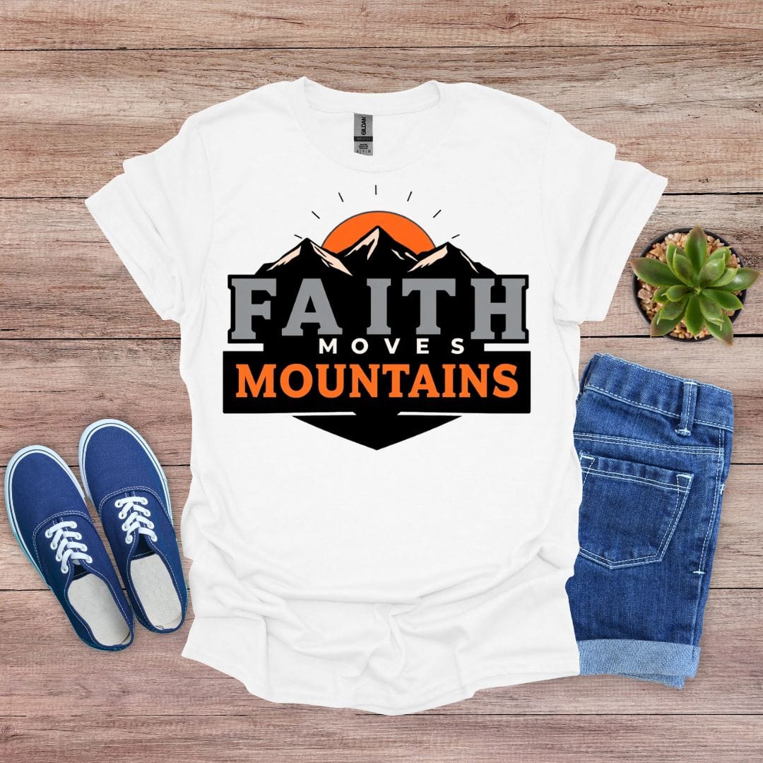 White t-shirt with 'Faith Moves Mountains' design on a wooden surface with blue shoes and jeans.