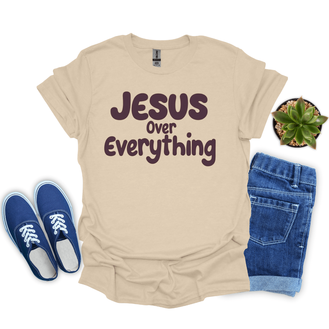 Sand t-shirt with 'Jesus Over Everything' text, blue sneakers, and jeans on a white background