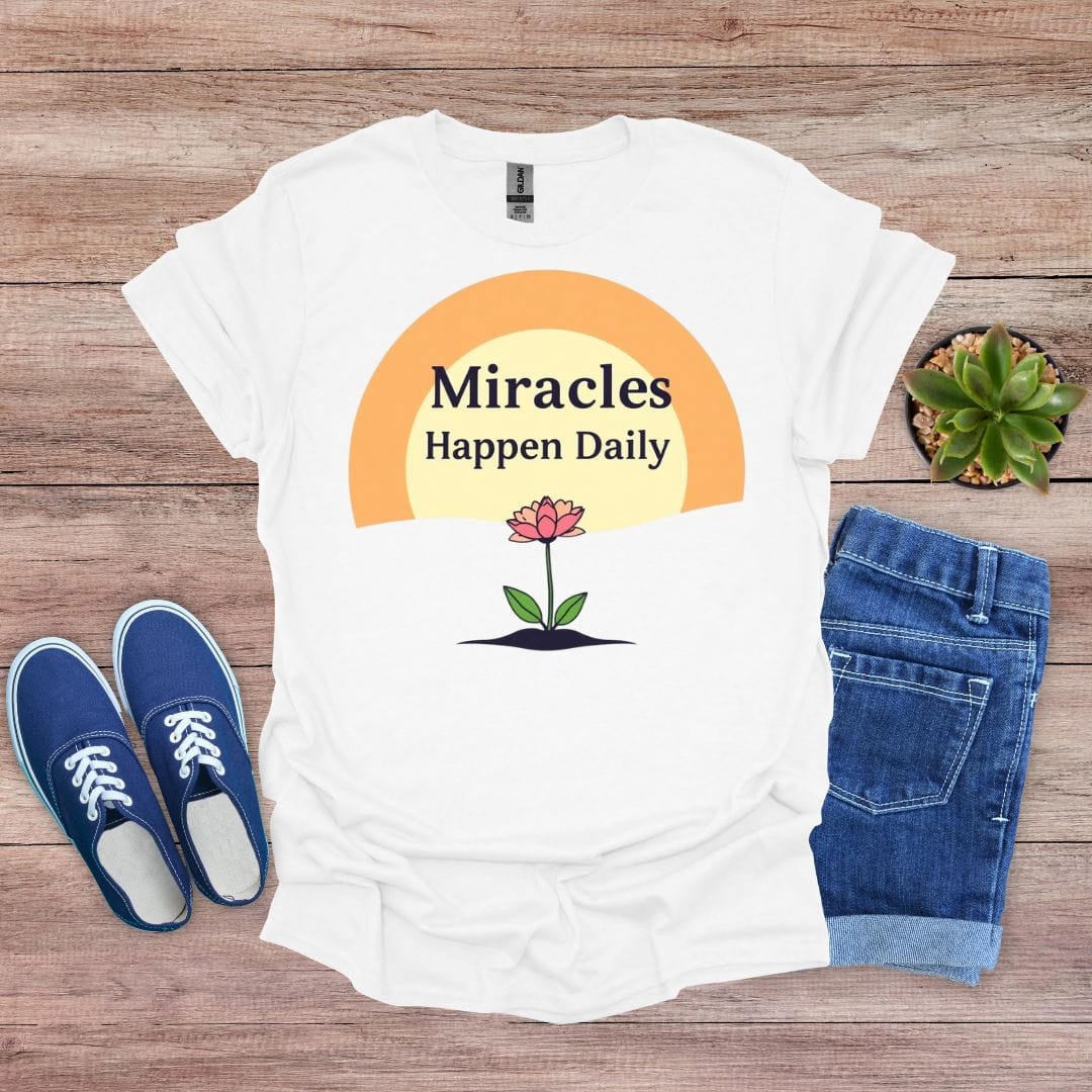 White t-shirt with 'Miracles Happen Daily' design on a wooden surface with shoes and jeans.