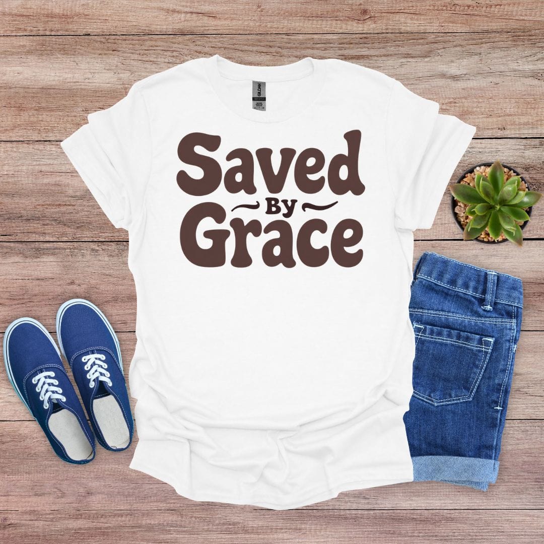 White t-shirt with 'Saved by Grace' text, blue shoes, and jeans on a wooden surface
